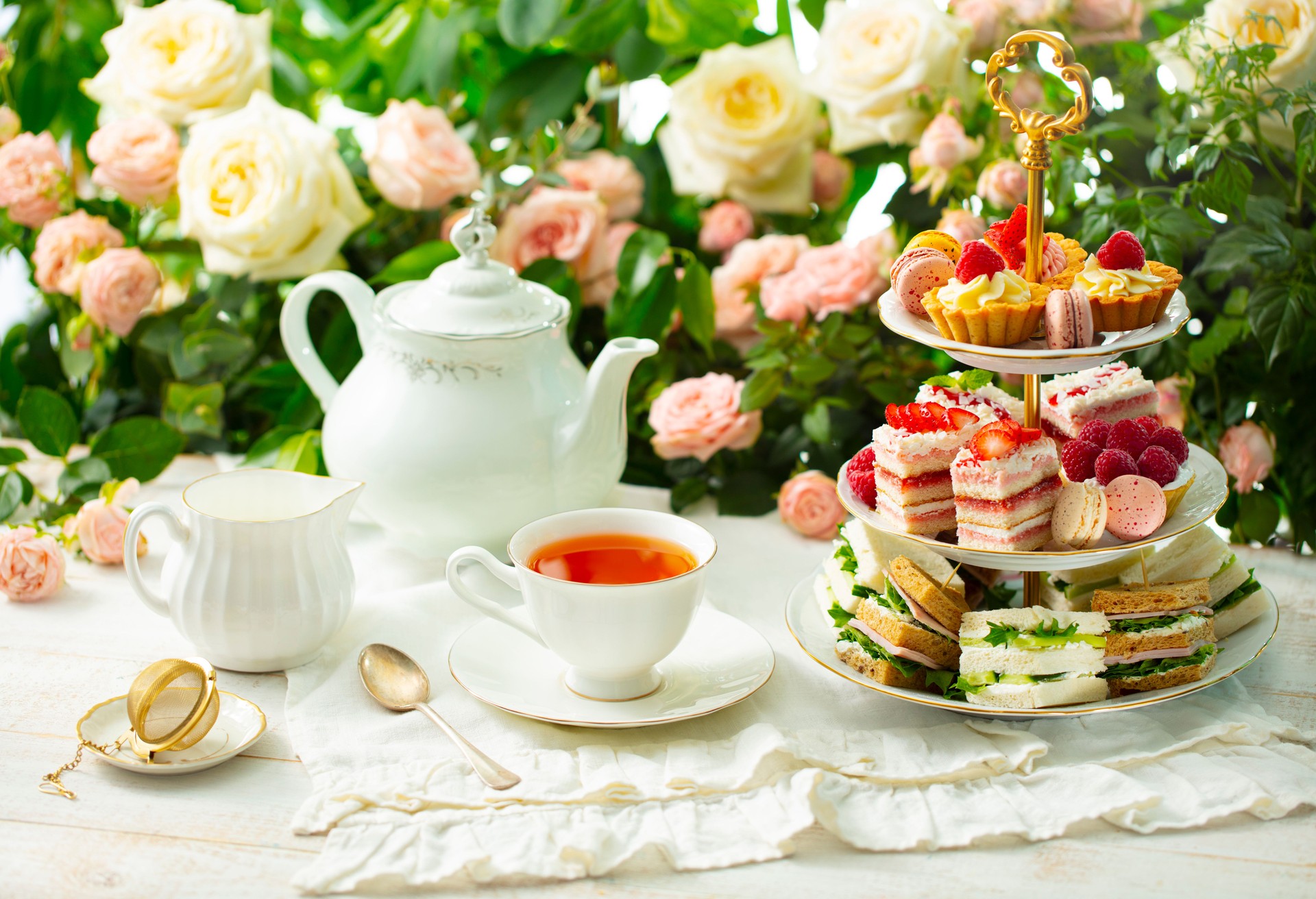 Afternoon tea table. Assortment of cakes, sweets and sandwiches. Rose garden background.